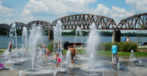 Children play among the water fountain feature with the scenic bridge view overlooking the riverfront. 